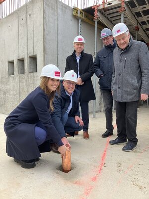 Bild vergr&ouml;&szlig;ern: F&uuml;nf Personen stehen auf der Baustelle um ein kleines Loch, in das zwei Personen ein Kupferrohr versenken.