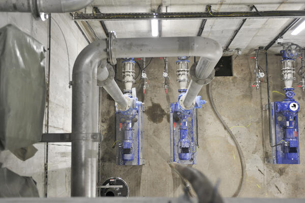 Bild vergrößern: In einem schlichten Pumpenraum befinden sich zwei große, blaue Pumpen, sowieein großeses Rohr, das die zwei Pumpen verbindet und nach oben steigt.