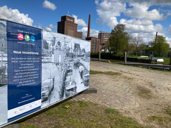 Bild vergrößern: Ein an einem Bauzaun befestigtes Großbanner mit einem historischen schwarz-weiß-Foto des Hafens steht am Nordufer des Elmshorner Hafens. Im Hintergrund sind die Köllnflocken-Werke zu sehen.