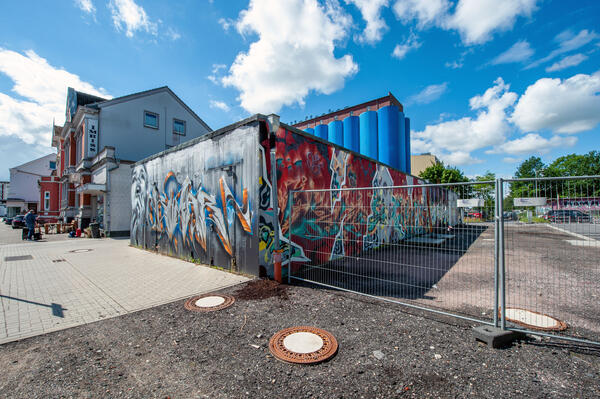 Bild vergrößern: Ein eingeschossiges, mit zahlreichen Graffiti verziertes Flachdach-Gebäude steht auf einer noch unbefestigten Fläche. Ein gründerzeitliches Gebäude sowie die blauen Silos der Kölln-Werke im Hintergrund flankieren den Rohbau.