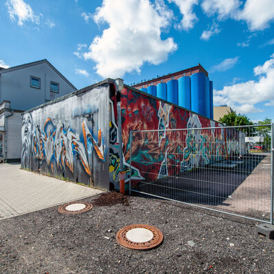 Ein eingeschossiges, mit zahlreichen Graffiti verziertes Flachdach-Gebäude steht auf einer noch unbefestigten Fläche. Ein gründerzeitliches Gebäude sowie die blauen Silos der Kölln-Werke im Hintergrund flankieren den Rohbau. 