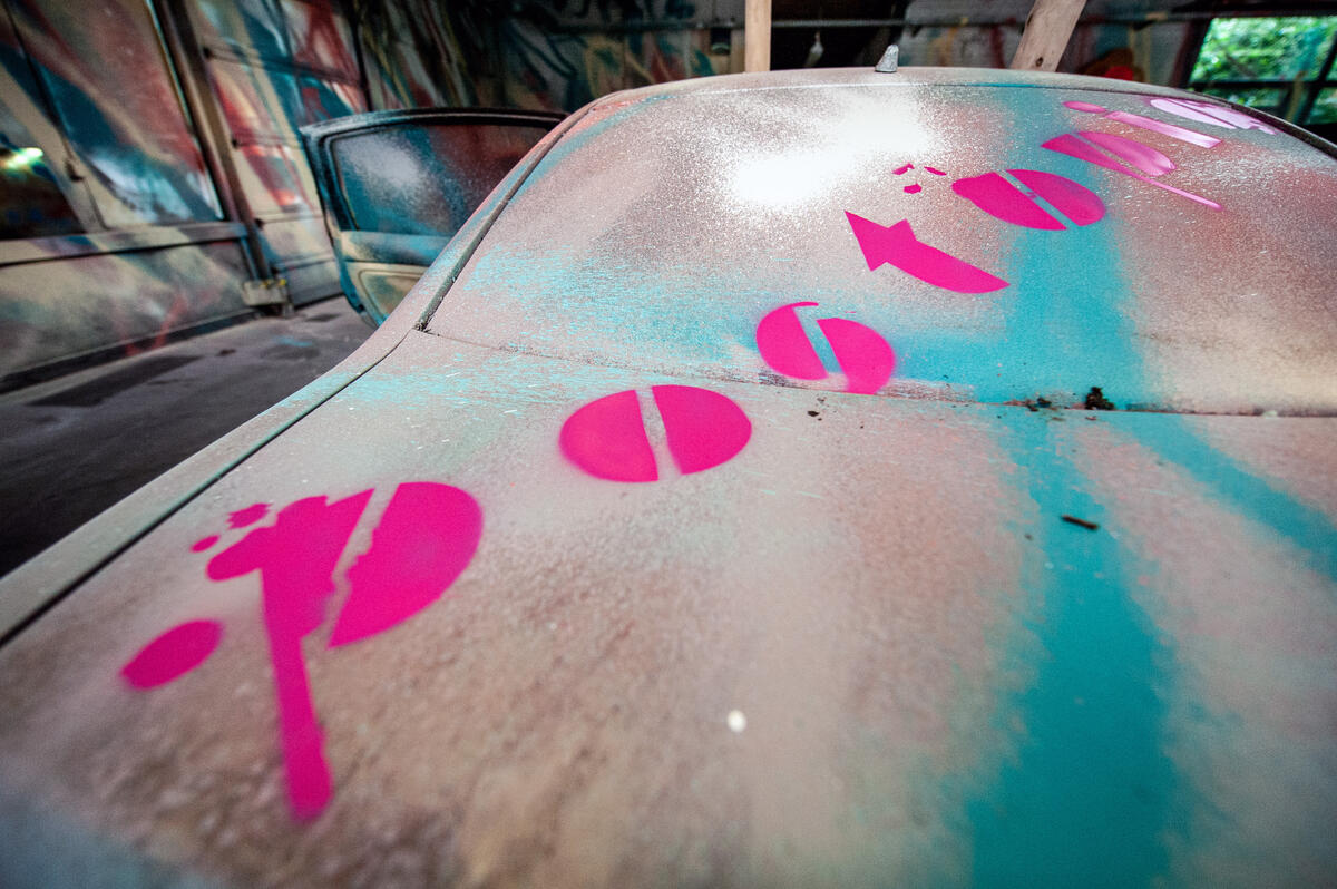 Bild vergrößern: Ein altes Auto steht in einer Halle. Sowohl das Auto, als auch die Halle wurden großflächig abstrakt-farbig gestaltet. Der Slogan "Postopia" prangt auf dem Kofferraum und der Heckscheibe des Autos.